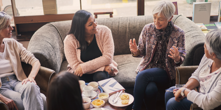 Women gathered in a cozy sitting area for a church women's ministry Bible study, with an open book and coffee on the table as an older woman leads the conversation.