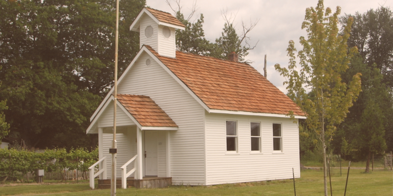 Small white country church with a cedar shake roof and steeple surrounded by trees, representing Christian education and discipleship in the local church.