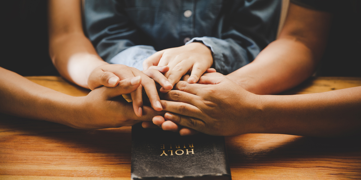 A parent and child holding hands over a Holy Bible, illustrating the importance of parents talking with their kids about the identity of Jesus.