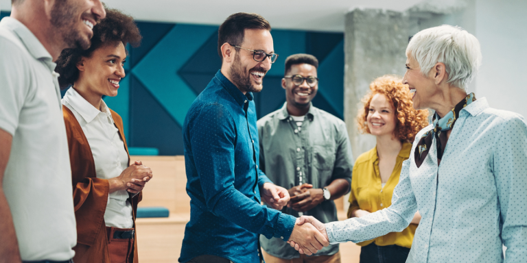 A diverse, multigenerational group of people smiling warmly as two individuals shake hands in greeting — illustrating the gospel-centered hospitality and genuine welcome that Matthew D. Kim calls churches to practice in Becoming a Friendlier Church: A Pathway to Genuine Community, published by Baker Books